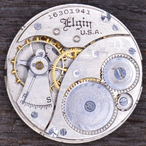 Puede incluir: Primer plano de un movimiento de reloj plateado con engranajes dorados. El reloj est&aacute; etiquetado como "Elgin U.S.A." y tiene el n&uacute;mero de serie "16301941".