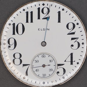 Op de afbeelding: Een wit zakhorloge-horloge met zwarte Romeinse cijfers voor de uren en zwarte Arabische cijfers voor de minuten. De wijzerplaat heeft een kleinere, witte sub-wijzerplaat met zwarte markeringen en een zwarte wijzer. De wijzerplaat heeft ook het woord "ELGIN" in het zwart gedrukt.