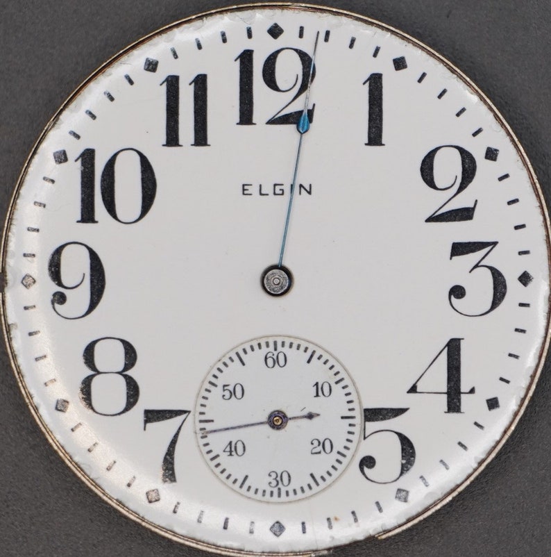 Op de afbeelding: Een wit zakhorloge-horloge met zwarte Romeinse cijfers voor de uren en zwarte Arabische cijfers voor de minuten. De wijzerplaat heeft een kleinere, witte sub-wijzerplaat met zwarte markeringen en een zwarte wijzer. De wijzerplaat heeft ook het woord "ELGIN" in het zwart gedrukt.
