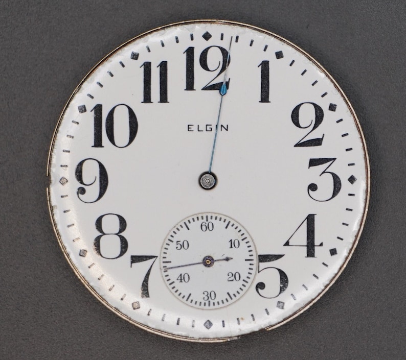 Op de afbeelding: Een wit zakhorloge met zwarte Romeinse cijfers voor de uren en zwarte Arabische cijfers voor de minuten. De wijzerplaat van het horloge heeft een kleine, ronde, witte subwijzerplaat met zwarte markeringen. De wijzerplaat van het horloge heeft ook het woord "ELGIN" erop gedrukt.