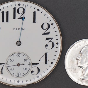 Op de afbeelding: Een wit zakhorloge-horloge met zwarte Romeinse cijfers voor de uren en zwarte Arabische cijfers voor de minuten. De wijzerplaat van het horloge heeft ook een kleinere, ronde sub-wijzerplaat met zwarte Arabische cijfers. De wijzerplaat van het horloge zegt "ELGIN" in het midden. Het horloge ligt naast een zilveren Amerikaanse kwartje.