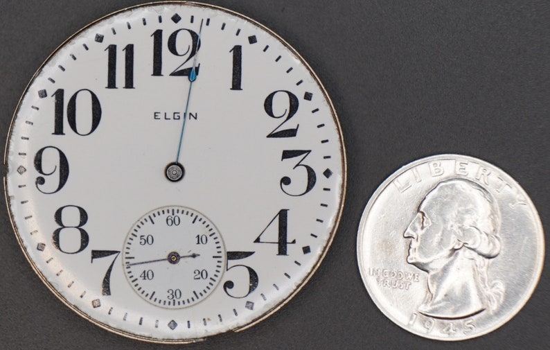Op de afbeelding: Een wit zakhorloge-horloge met zwarte Romeinse cijfers voor de uren en zwarte Arabische cijfers voor de minuten. De wijzerplaat van het horloge heeft ook een kleinere, ronde sub-wijzerplaat met zwarte Arabische cijfers. De wijzerplaat van het horloge zegt "ELGIN" in het midden. Het horloge ligt naast een zilveren Amerikaanse kwartje.