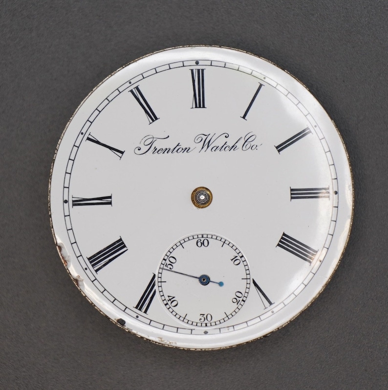 May include: White pocket watch face with black Roman numeral hour markers and black markings for the seconds. The watch face has the text "Trenton Watch Co." printed on it.