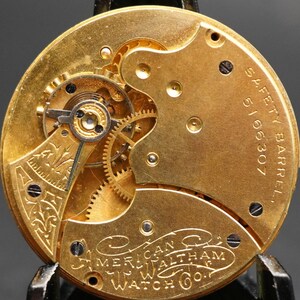 Puede incluir: Primer plano de un movimiento de reloj de bolsillo dorado con engranajes intrincados y un barril de seguridad marcado con "5166307". El reloj est&aacute; inscrito con "American Waltham Watch Co."