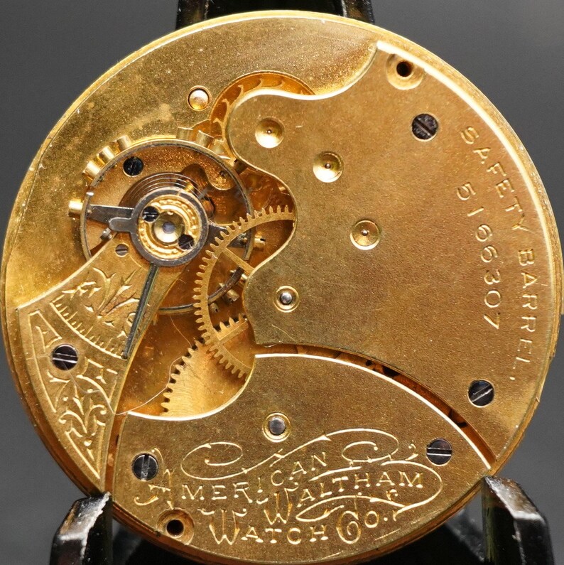Puede incluir: Primer plano de un movimiento de reloj de bolsillo dorado con engranajes intrincados y un barril de seguridad marcado con "5166307". El reloj est&aacute; inscrito con "American Waltham Watch Co."