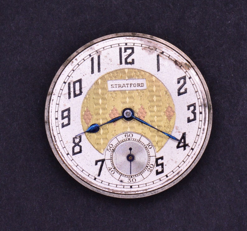 Pu&ograve; includere: Un orologio da tasca argentato con un quadrante bianco e indici orari romani neri. L'orologio ha un cerchio interno dorato con un motivo a rombi e la parola "STRATFORD" stampata in nero. L'orologio ha una lancetta blu e un quadrante bianco pi&ugrave; piccolo con segni neri per i secondi.
