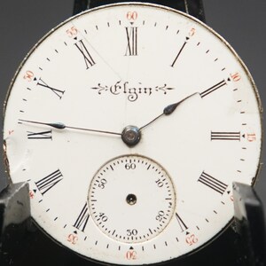 Puede incluir: Una esfera de reloj de bolsillo blanca con marcadores de hora romanos negros y agujas negras. La esfera del reloj tiene un c&iacute;rculo conc&eacute;ntrico m&aacute;s peque&ntilde;o con marcas negras para los segundos. La esfera del reloj est&aacute; etiquetada como "Elgin" en una escritura cursiva.