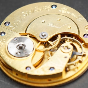 May include: Close-up view of the inner workings of a gold-toned pocket watch. The watch face is engraved with the text " Elgin National Watch Co. Dexter St.  Patented".