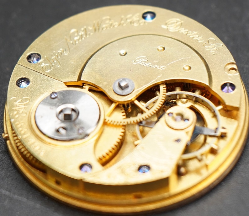 May include: Close-up view of the inner workings of a gold-toned pocket watch. The watch face is engraved with the text " Elgin National Watch Co. Dexter St.  Patented".