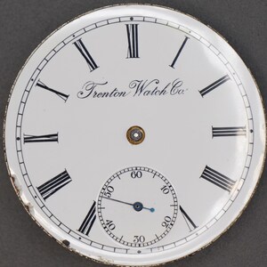 May include: White pocket watch face with black Roman numeral hour markers and black minute markers. The watch face has a smaller, white sub-dial with black minute markers and a blue hand. The watch face also has the text "Trenton Watch Co."