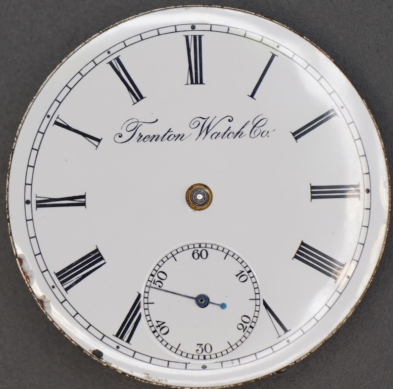 May include: White pocket watch face with black Roman numeral hour markers and black minute markers. The watch face has a smaller, white sub-dial with black minute markers and a blue hand. The watch face also has the text "Trenton Watch Co."