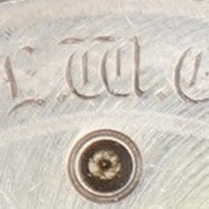 以下が含まれることがあります： 「T.L.C. Co.」という刻印がされた銀色の時計の文字盤のクローズアップ
