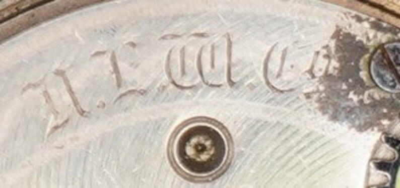 以下が含まれることがあります： 「T.L.C. Co.」という刻印がされた銀色の時計の文字盤のクローズアップ