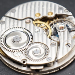 May include: Close-up of a silver pocket watch movement with intricate gears and a silver dial. The watch face is visible with the text "17 Jewels" printed on it.