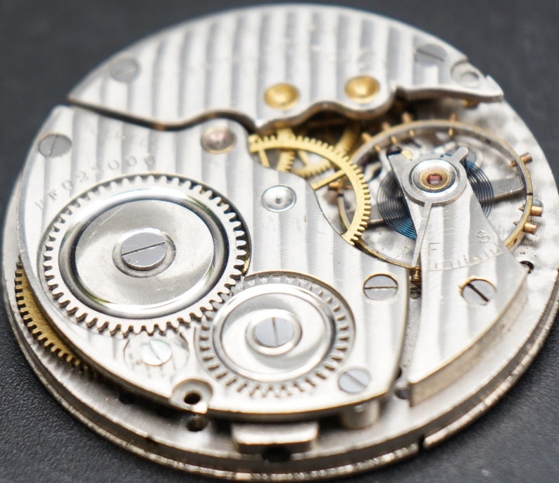 May include: Close-up of a silver pocket watch movement with intricate gears and a silver dial. The watch face is visible with the text "17 Jewels" printed on it.