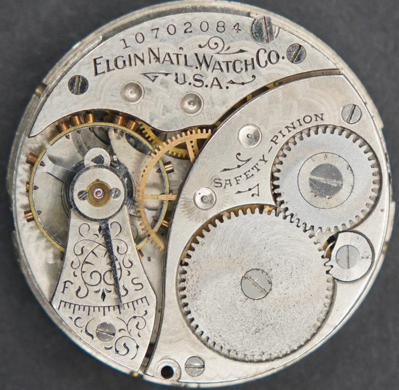Puede incluir: Primer plano de un movimiento de reloj de bolsillo plateado con el texto "ELGIN NAT'L WATCH CO. U.S.A." y "SAFETY-PINION" visible. El movimiento del reloj tiene una serie de engranajes y un v&aacute;stago de cuerda.