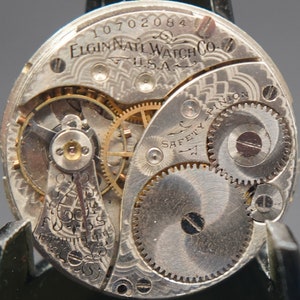 Puede incluir: Primer plano de un movimiento de reloj de bolsillo plateado con la inscripci&oacute;n "Elgin National Watch Co. U.S.A." y el n&uacute;mero "10702084". El movimiento presenta engranajes intrincados y un pasador de seguridad.