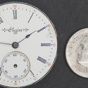 Puede incluir: Una esfera de reloj de bolsillo blanca con marcadores de hora en n&uacute;meros romanos negros y agujas negras. La esfera del reloj tiene la palabra "Elgin" impresa en escritura negra. La esfera del reloj es aproximadamente del tama&ntilde;o de una moneda de diez c&eacute;ntimos de euro.