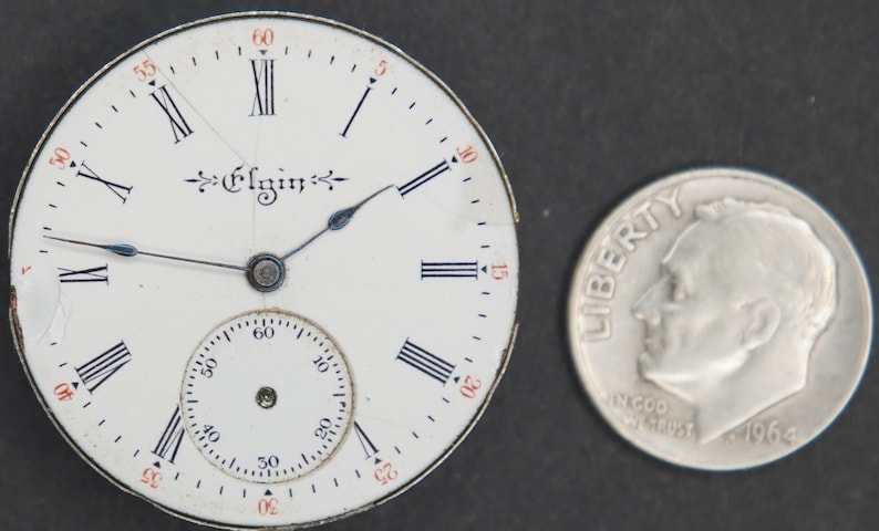 Puede incluir: Una esfera de reloj de bolsillo blanca con marcadores de hora en n&uacute;meros romanos negros y agujas negras. La esfera del reloj tiene la palabra "Elgin" impresa en escritura negra. La esfera del reloj es aproximadamente del tama&ntilde;o de una moneda de diez c&eacute;ntimos de euro.