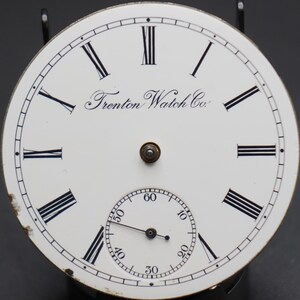 May include: White watch face with black Roman numeral hour markers and black minute markers. The watch face has the text "Trenton Watch Co." printed on it.