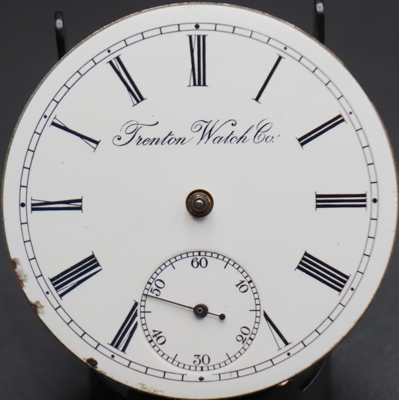May include: White watch face with black Roman numeral hour markers and black minute markers. The watch face has the text "Trenton Watch Co." printed on it.