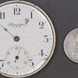 May include: A white pocket watch face with black Roman numeral hour markers and a smaller, concentric dial with black markings. The watch face is cracked and the glass is broken. The watch face has the text "Standard U.S.A." printed on it.