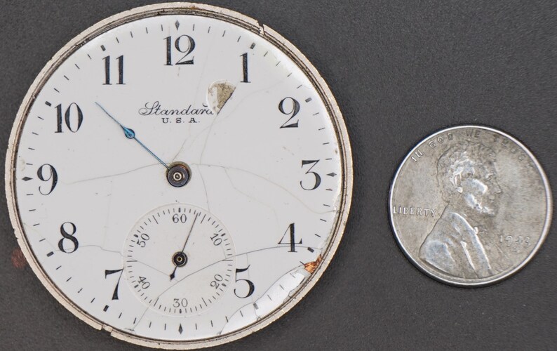 May include: A white pocket watch face with black Roman numeral hour markers and a smaller, concentric dial with black markings. The watch face is cracked and the glass is broken. The watch face has the text "Standard U.S.A." printed on it.
