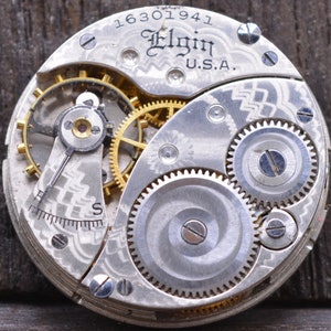 Puede incluir: Primer plano de un movimiento de reloj de bolsillo plateado con engranajes intrincados y la inscripci&oacute;n "Elgin U.S.A." y el n&uacute;mero "16301941".