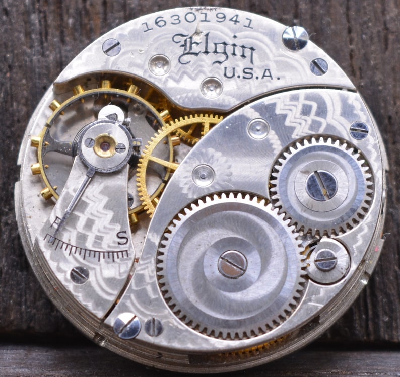 Puede incluir: Primer plano de un movimiento de reloj de bolsillo plateado con engranajes intrincados y la inscripci&oacute;n "Elgin U.S.A." y el n&uacute;mero "16301941".