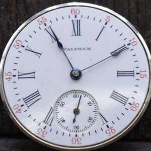 May include: A white pocket watch with Roman numeral markings and a smaller sub-dial. The watch face says "Waltham" and has black hands.