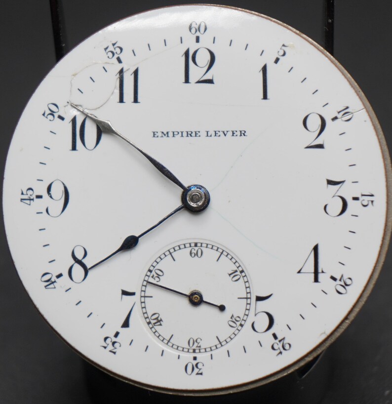 May include: A white pocket watch face with black Roman numeral hour markers and black hands. The watch face has a small sub-dial at the bottom and the text "EMPIRE LEVER" printed in black.