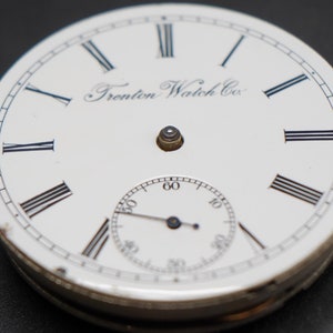 May include: White pocket watch face with black Roman numeral hour markers and black hands. The watch face has the text "Trenton Watch Co." printed on it.