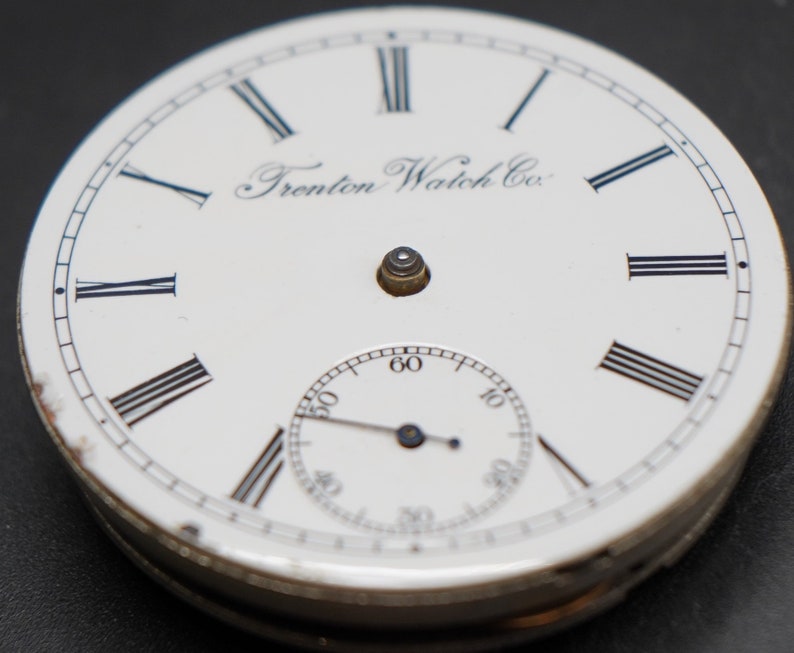 May include: White pocket watch face with black Roman numeral hour markers and black hands. The watch face has the text "Trenton Watch Co." printed on it.