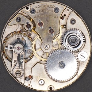 May include: Close-up view of a silver watch movement with intricate gears and the text "ADJUSTED 166-212 15 FIFTEEN JEWELS".