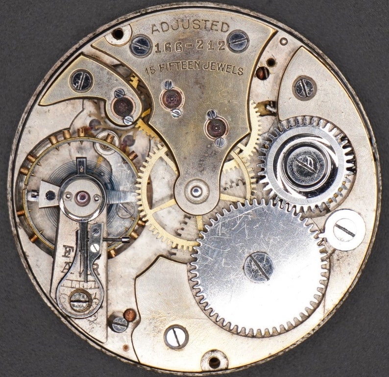 May include: Close-up view of a silver watch movement with intricate gears and the text "ADJUSTED 166-212 15 FIFTEEN JEWELS".