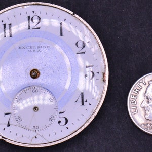 May include: A white watch face with black numbers and hands. The watch face is cracked and has a blue ring around the edge. The watch face says "EXCELSIOR USA" in the center. A dime is next to the watch face for scale.