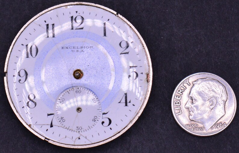 May include: A white watch face with black numbers and hands. The watch face is cracked and has a blue ring around the edge. The watch face says "EXCELSIOR USA" in the center. A dime is next to the watch face for scale.