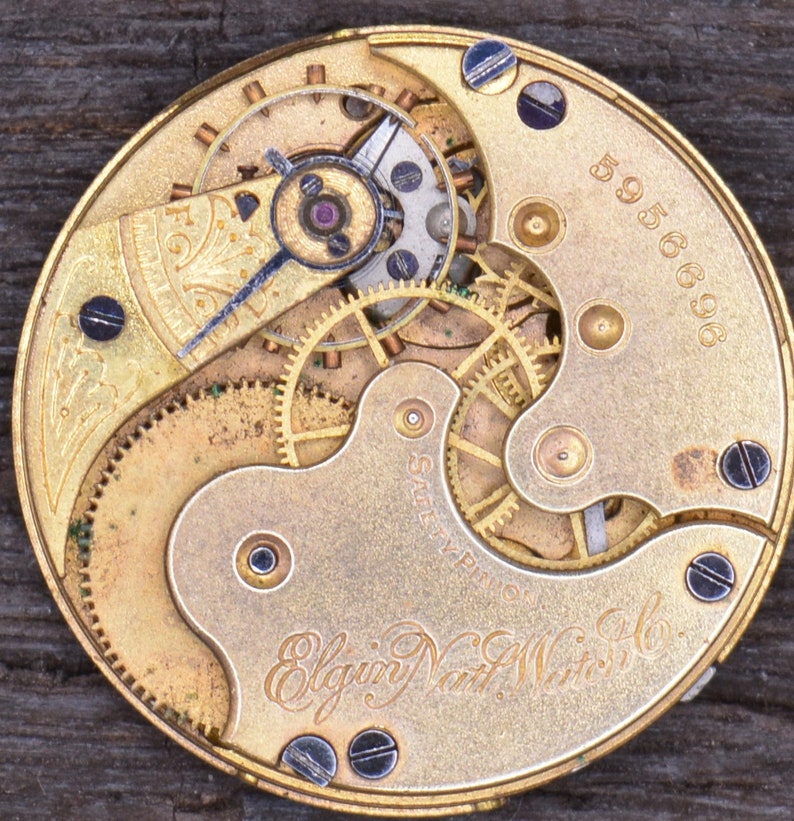 May include: Close-up of a gold-toned pocket watch movement with intricate gears and a '5956696' serial number. The watch is labelled 'Elgin Natl. Watch Co.'