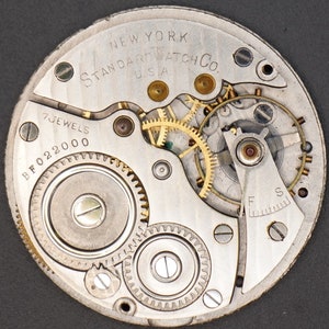 May include: Close-up of a silver watch movement with gold gears. The watch is labeled "New York Standard Watch Co. U.S.A." and "7 Jewels BF022000".