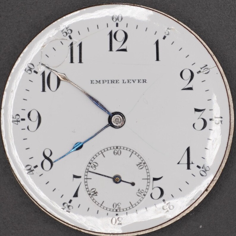 May include: A white pocket watch face with black Roman numeral hour markers and black Arabic numeral minute markers. The watch face has a small seconds dial at the bottom. The watch face says "EMPIRE LEVER".