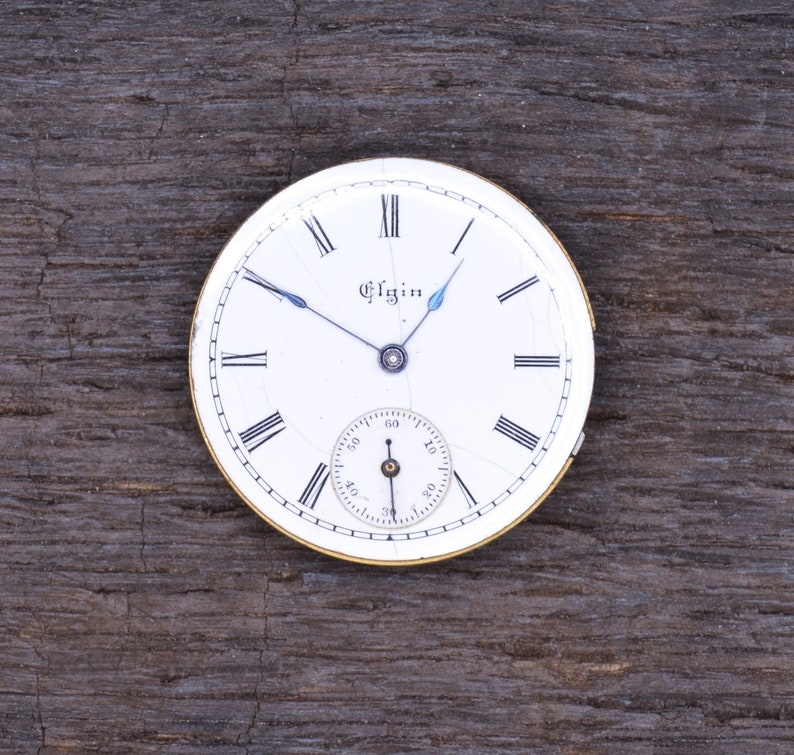 May include: A white pocket watch face with black Roman numeral hour markers and black hands. The watch face has a small seconds dial and the word "Elgin" printed in black.