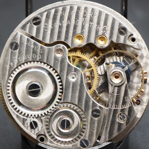 May include: Close-up view of the inner workings of a silver pocket watch. The watch face is marked "NEW YORK STANDARD WATCH CO. U.S.A." and "17 JEWELS BFO 2200".