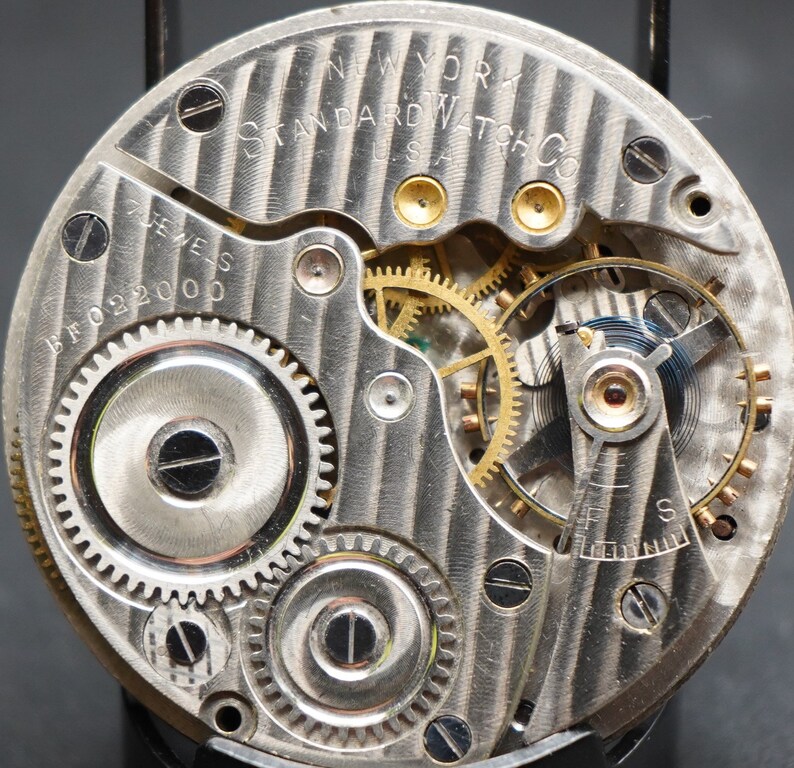 May include: Close-up view of the inner workings of a silver pocket watch. The watch face is marked "NEW YORK STANDARD WATCH CO. U.S.A." and "17 JEWELS BFO 2200".