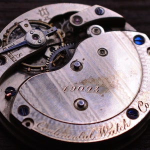 May include: Close-up of the back of a silver pocket watch with the inscription "Continental Watch Co." and the number "19093". The watch has a complex gear system visible.