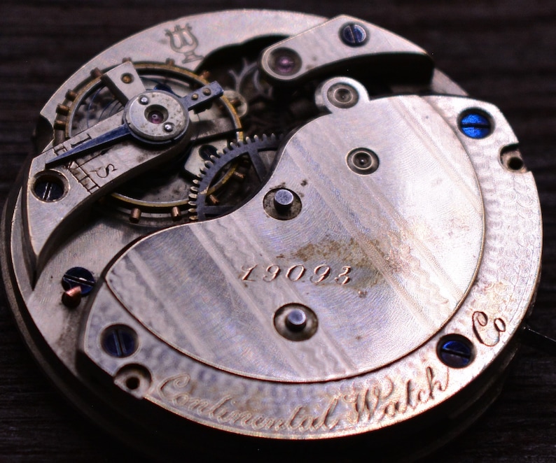 May include: Close-up of the back of a silver pocket watch with the inscription "Continental Watch Co." and the number "19093". The watch has a complex gear system visible.