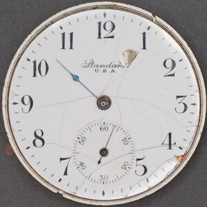 May include: A white pocket watch with black Roman numeral hour markers and black Arabic numeral minute markers. The watch face is cracked and has a small, round, white sub-dial with black markings. The watch face also has the text "Standard U.S.A." printed on it.