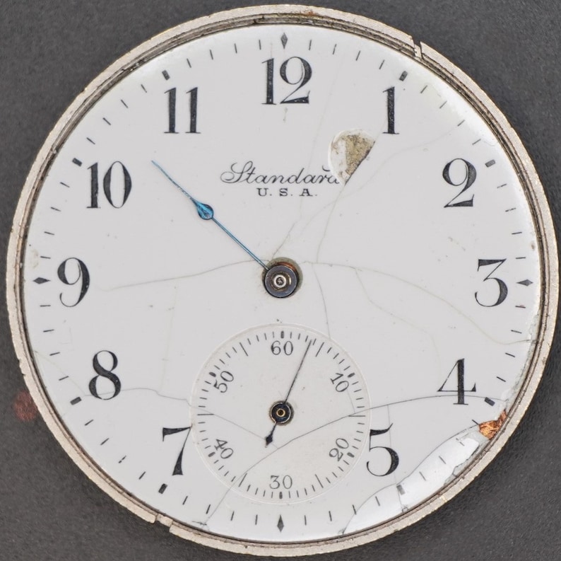 May include: A white pocket watch with black Roman numeral hour markers and black Arabic numeral minute markers. The watch face is cracked and has a small, round, white sub-dial with black markings. The watch face also has the text "Standard U.S.A." printed on it.