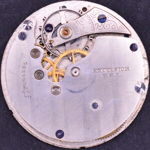 May include: Close-up of the back of a silver pocket watch with a visible gear mechanism. The watch face has the text "EXCELSIOR U.S.A." and the number "A2W015285".