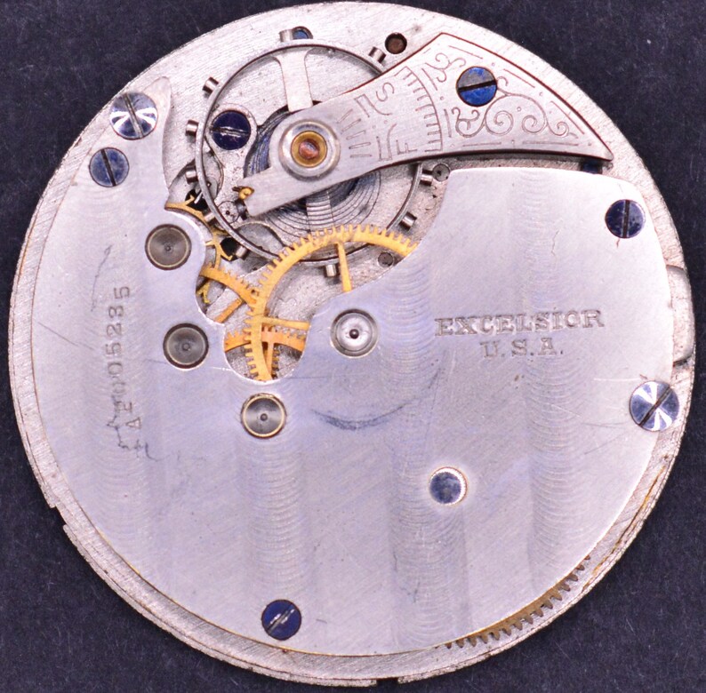 May include: Close-up of the back of a silver pocket watch with a visible gear mechanism. The watch face has the text "EXCELSIOR U.S.A." and the number "A2W015285".