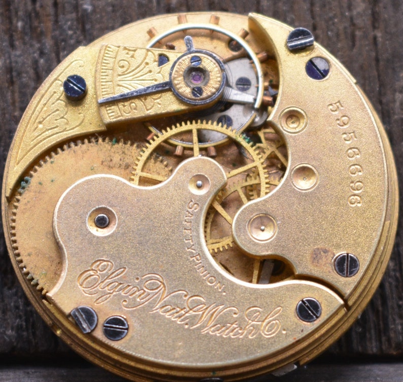 May include: Close-up of a gold-toned pocket watch movement with intricate gears and an 'Elgin Natl. Watch Co.' inscription. The watch has a serial number '5956696'.